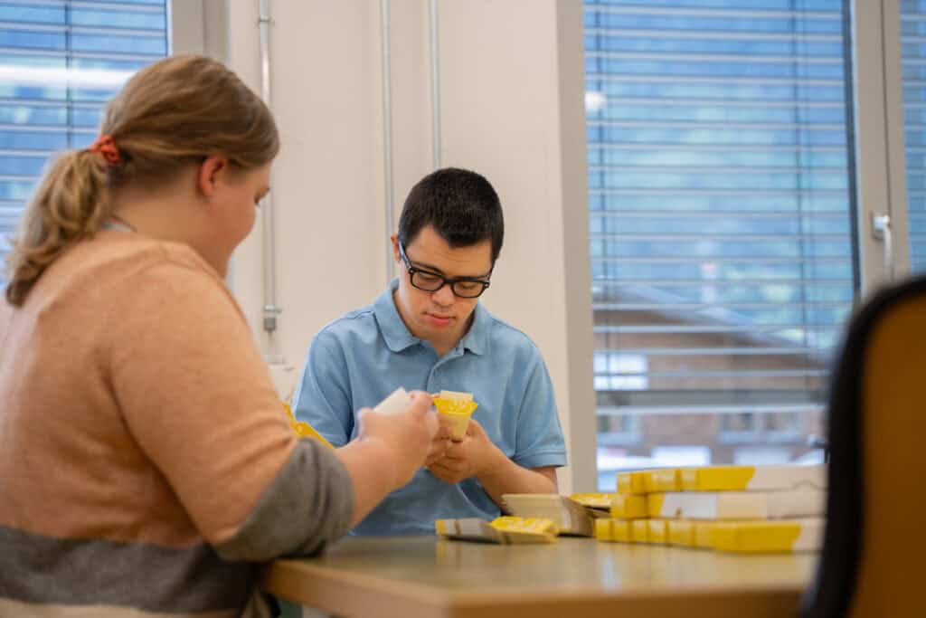 Zwei Personen sitzen an einem Tisch und bauen gelbe Kartons zusammen. Eine Person, die ein blaues Hemd und eine Brille trägt, ist konzentriert auf die Aufgabe. Die andere Person ist abgewandt und trägt einen beigen Pullover. Im Hintergrund sind große Fenster mit Jalousien zu sehen.