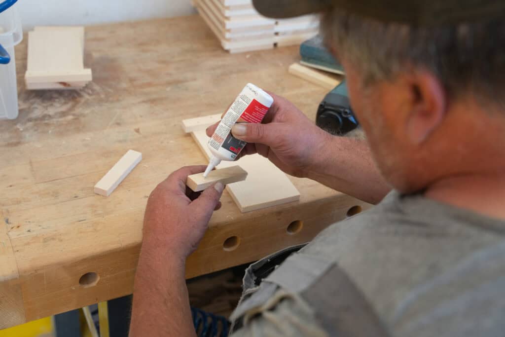 Eine Person trägt in einer Werkstatt Klebstoff auf ein kleines Holzstück auf. In der Nähe liegen weitere Holzstücke auf einer Werkbank. Sie trägt ein graues Hemd und eine Mütze und konzentriert sich darauf, das Holz zusammenzusetzen.