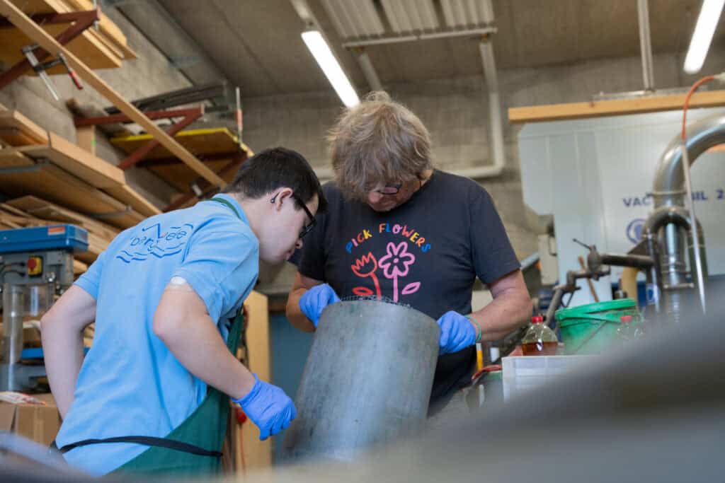 Zwei Personen mit Handschuhen arbeiten gemeinsam in einer Werkstatt. Die ältere Person, in einem schwarzen Hemd mit Blumenmuster, hält ein großes Metallobjekt. Die jüngere Person, in einem blauen Hemd, beugt sich vor, um zuzusehen. Im Hintergrund sind Werkzeuge und Holzregale zu sehen.