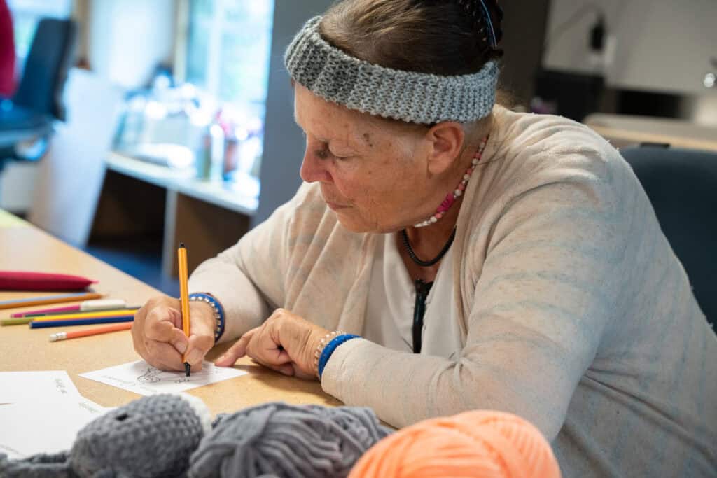 Eine ältere Frau mit grauem Stirnband sitzt an einem Tisch und zeichnet mit einem Bleistift. Um sie herum liegen Buntstifte, Garn und Bastelmaterialien verstreut. Sie ist konzentriert in einem gut beleuchteten Raum mit Fenstern im Hintergrund.