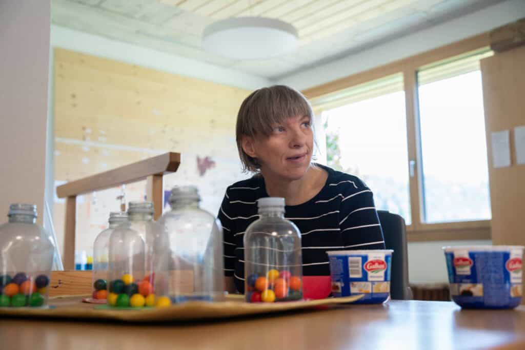 Eine Person mit kurzen Haaren sitzt an einem Tisch mit verschiedenen Gegenständen, darunter Plastikflaschen, bunte Bonbons und Joghurtbecher. Der Raum hat Holzwände und ein Fenster, durch das natürliches Licht hereinfällt.