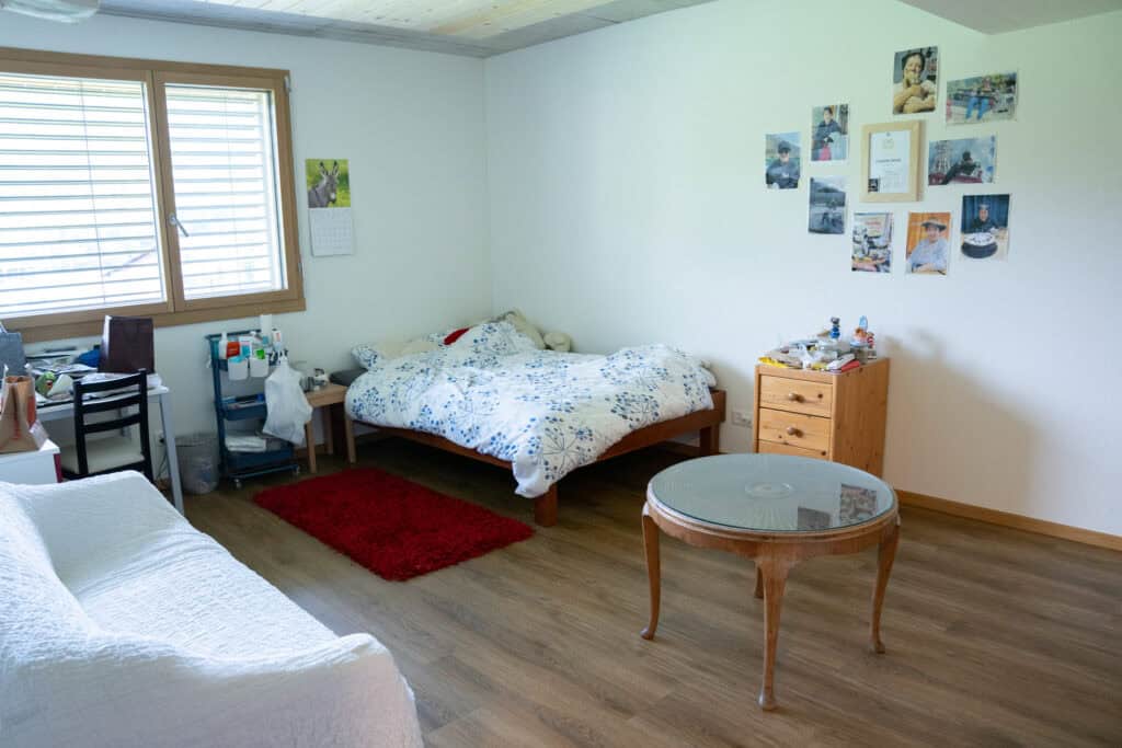 Ein gemütliches Schlafzimmer mit einem Bett an der Wand, das mit einer weiß-blauen Bettdecke bedeckt ist. Daneben steht ein Nachttisch aus Holz mit Gegenständen. Im Vordergrund steht ein runder Couchtisch aus Glas, links ein Sofa und an der Wand ein Fenster mit Jalousien.