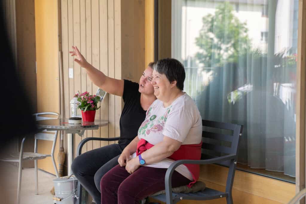 Zwei Frauen sitzen auf einer Bank auf einer Veranda. Eine Frau zeigt auf etwas in der Ferne und beide lächeln. In der Nähe steht ein kleiner Tisch mit einer Pflanze. Sie genießen einen entspannten Moment im Freien.
