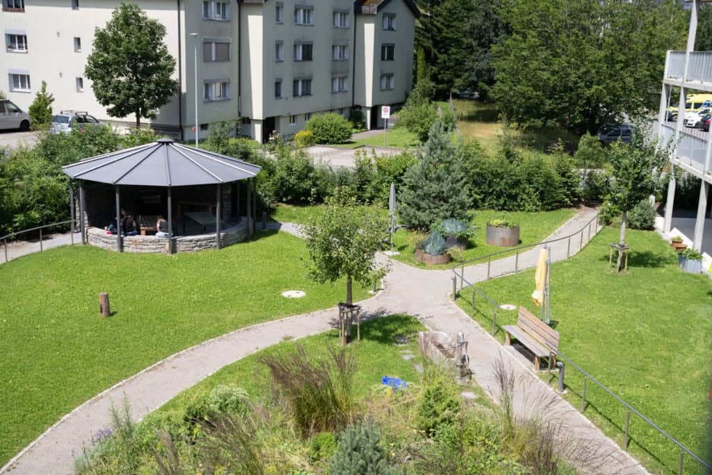 Luftaufnahme eines kleinen grünen Parks mit Spazierwegen, einem überdachten Steinpavillon, Bänken und einer Landschaftsgestaltung mit Bäumen und Sträuchern. Im Hintergrund sind Wohngebäude zu sehen.