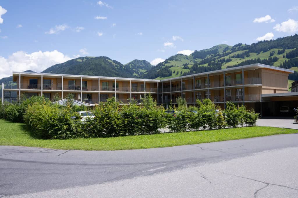 Ein modernes Holzgebäude mit großen Fenstern vor einer malerischen Bergkulisse unter blauem Himmel. Grünflächen und Hecken umgeben das Gebäude, im Vordergrund verläuft eine asphaltierte Straße.