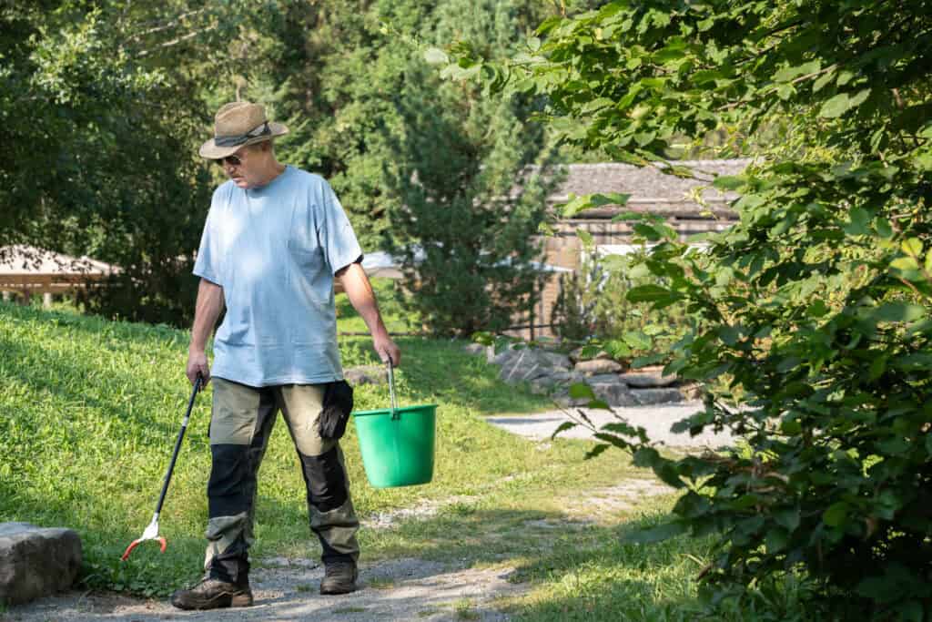 Eine Person mit Hut und Sonnenbrille verwendet ein Greifwerkzeug, um in einem Park Müll aufzusammeln. Sie hält einen grünen Eimer und geht an einem sonnigen Tag einen Kiesweg entlang, der von Bäumen und Gras umgeben ist.