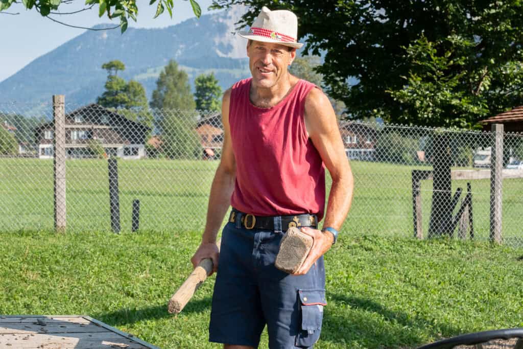Ein Mann mit einem roten Tanktop, Shorts und Hut steht im Freien und hält einen Hammer und einen Holzblock in der Hand. Hinter ihm sind eine Wiese, Bäume und eine Berglandschaft unter einem klaren blauen Himmel zu sehen.