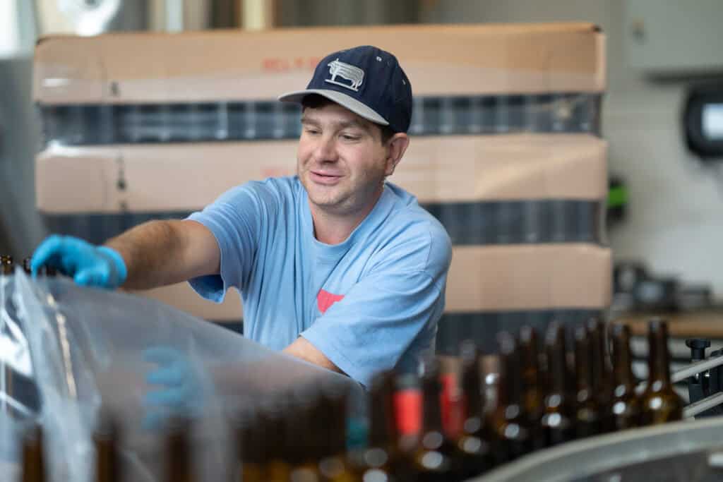 Eine Person mit blauer Mütze und Handschuhen packt in einer Brauerei Bierflaschen in eine Kiste. Sie lächelt und ist von Flaschenreihen umgeben. Im Hintergrund sind Kartons gestapelt.