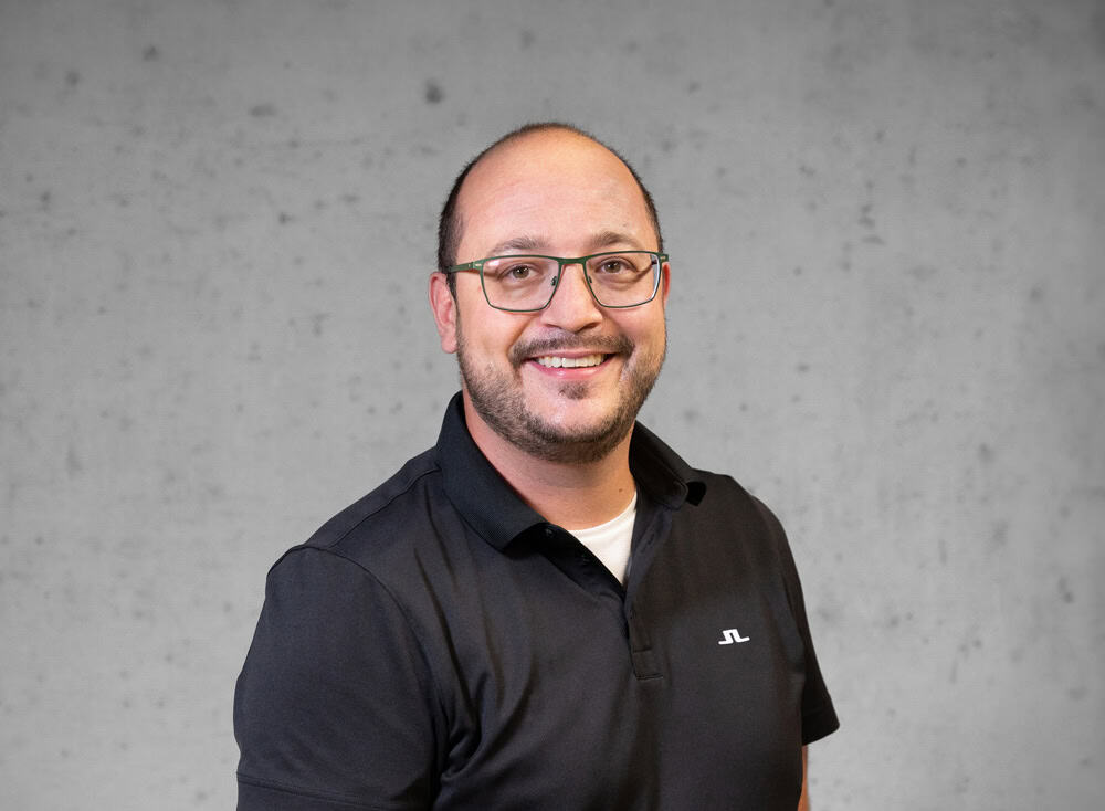 Ein Mann mit Brille und Bart lächelt in die Kamera und trägt ein schwarzes Poloshirt. Der Hintergrund ist eine schlichte graue Wand.