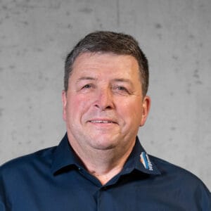 Ein Mann mit kurzen Haaren, der ein marineblaues Hemd mit Kragen und einem Logo auf der Schulter trägt, lächelt vor einem schlichten, strukturierten Hintergrund.