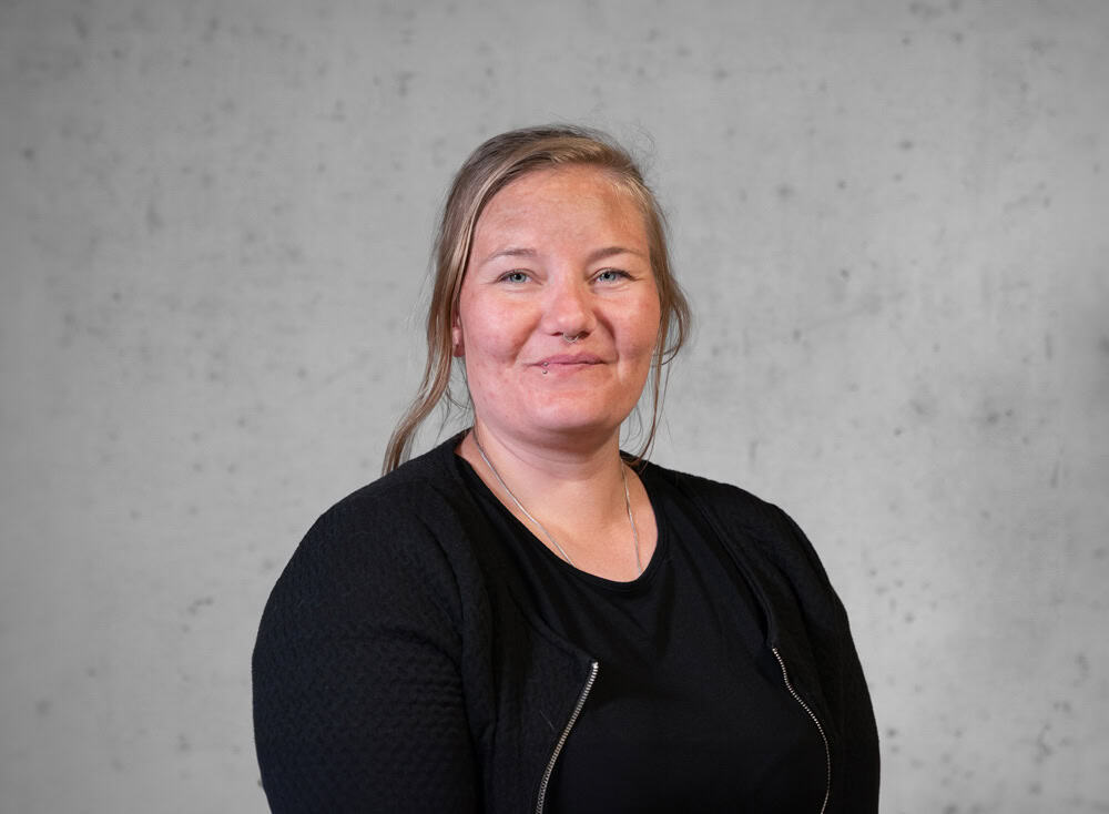Eine Person mit langen Haaren lächelt herzlich und trägt ein schwarzes Oberteil und eine Jacke. Der Hintergrund ist eine schlichte, helle Wand.