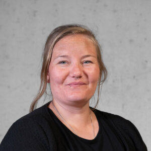 Eine Frau mit langen Haaren, einem schwarzen Top und einer Jacke, lächelt sanft in die Kamera. Sie hat ein Nasenpiercing und steht vor einem schlichten, hellen Hintergrund.