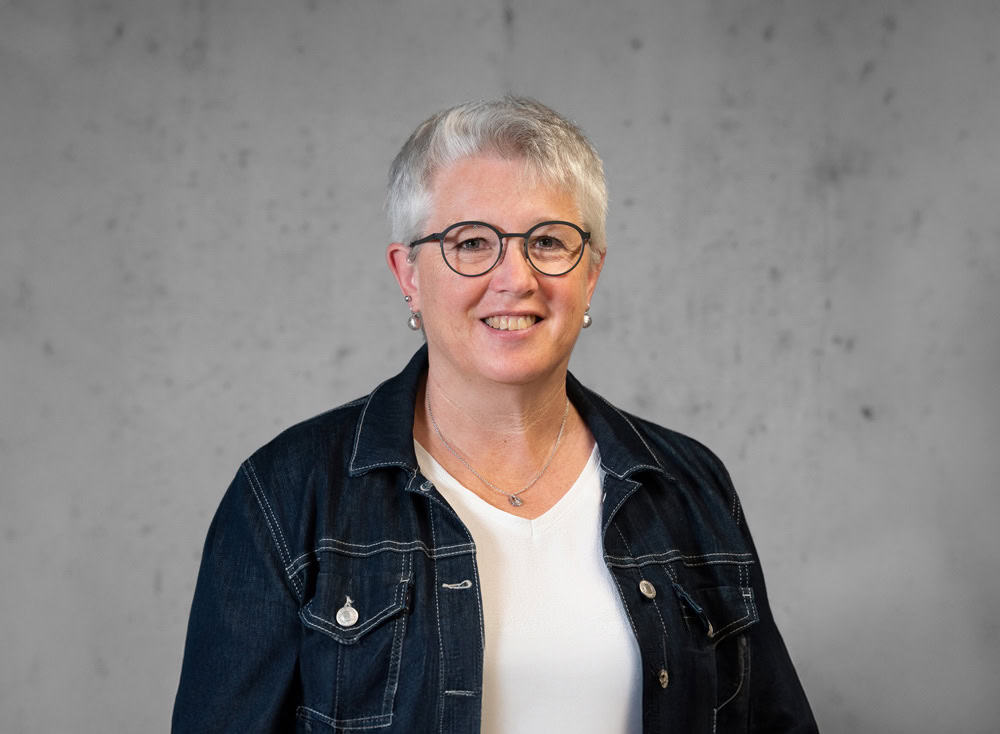 Eine Person mit kurzen grauen Haaren und Brille lächelt in die Kamera. Sie trägt eine Jeansjacke über einem weißen Hemd und steht vor einem schlichten grauen Hintergrund.