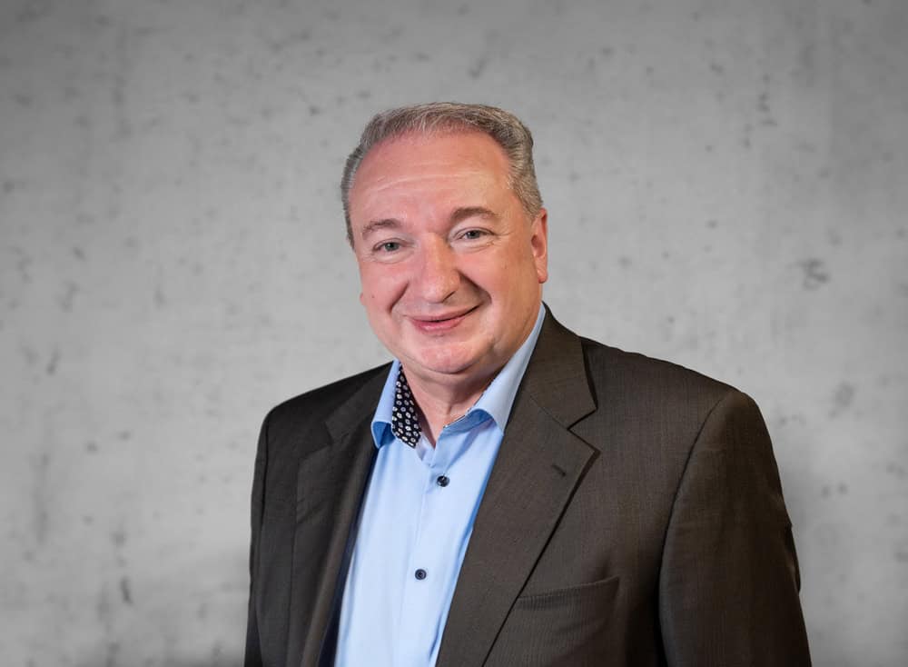 Ein lächelnder Mann mit kurzen, ergrauenden Haaren trägt ein dunkles Jackett über einem blauen Hemd mit Knöpfen. Der Hintergrund ist eine schlichte, helle Wand.