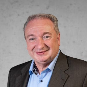 Lächelnde Person mit kurzen, grauen Haaren, bekleidet mit einem dunklen Jackett und einem hellblauen Hemd, steht vor einem gesprenkelten grauen Hintergrund.