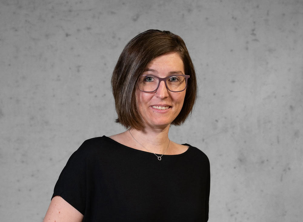 Eine Frau mit schulterlangem braunem Haar und Brille sowie einem schwarzen Hemd steht vor einer strukturierten grauen Wand. Sie lächelt in die Kamera.