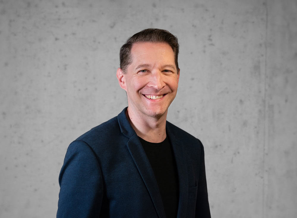 Eine Person mit kurzen braunen Haaren lächelt in die Kamera. Sie trägt einen dunklen Blazer über einem schwarzen Hemd. Der Hintergrund ist eine schlichte, strukturierte Wand.