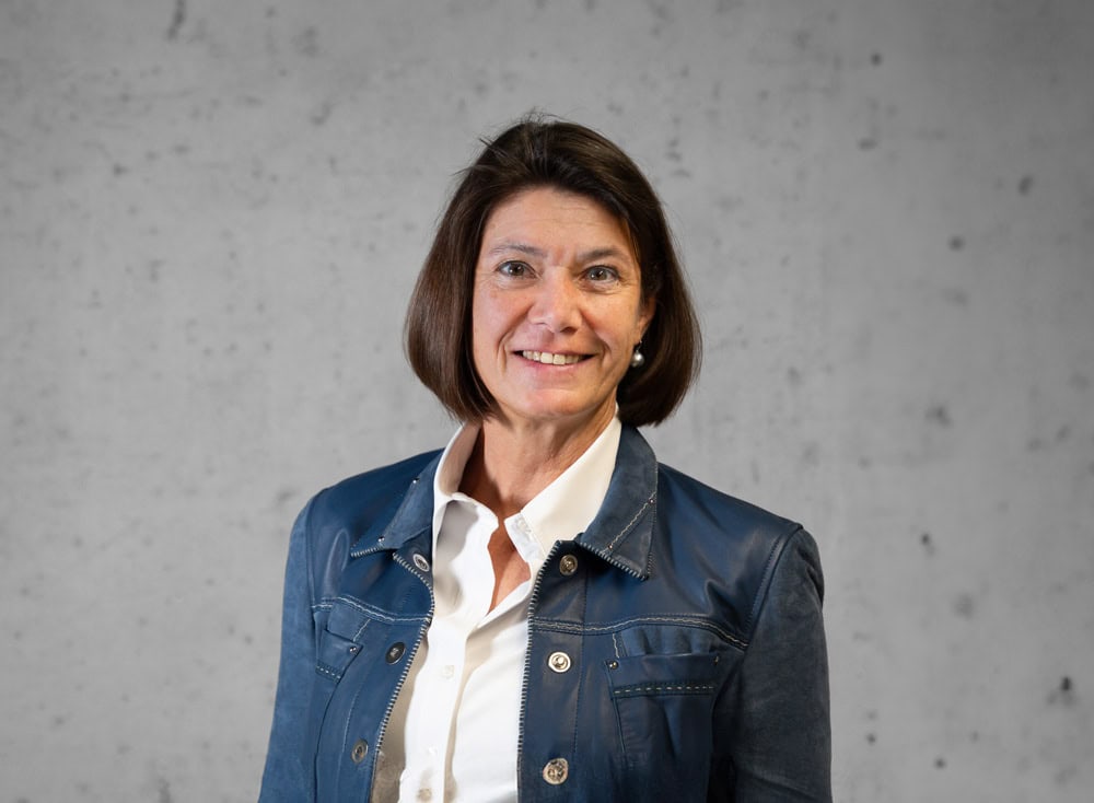 Eine Frau mit kurzen braunen Haaren lächelt in die Kamera. Sie trägt eine blaue Jacke über einem weißen Hemd. Der Hintergrund ist eine schlichte, strukturierte Wand.