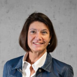 Eine Frau mit kurzen braunen Haaren lächelt und trägt eine blaue Jacke über einem weißen Hemd. Der Hintergrund ist eine schlichte graue Wand.