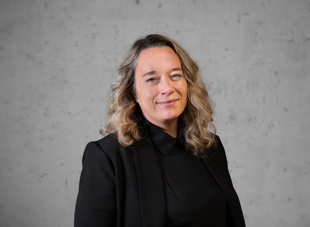 Eine Person mit schulterlangem, welligem Haar, einem schwarzen Blazer und Hemd steht vor einem schlichten grauen Hintergrund und lächelt sanft in die Kamera.
