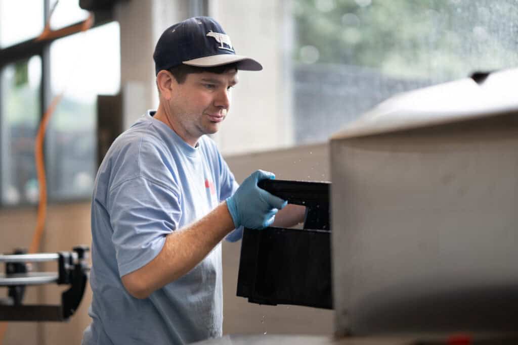 Ein Mann mit blauer Mütze und Handschuhen arbeitet an Maschinen in einer Industrieanlage. Er konzentriert sich auf seine Aufgabe und hält einen schwarzen Gegenstand in der Hand. Natürliches Licht aus einem Fenster erhellt den Arbeitsbereich.