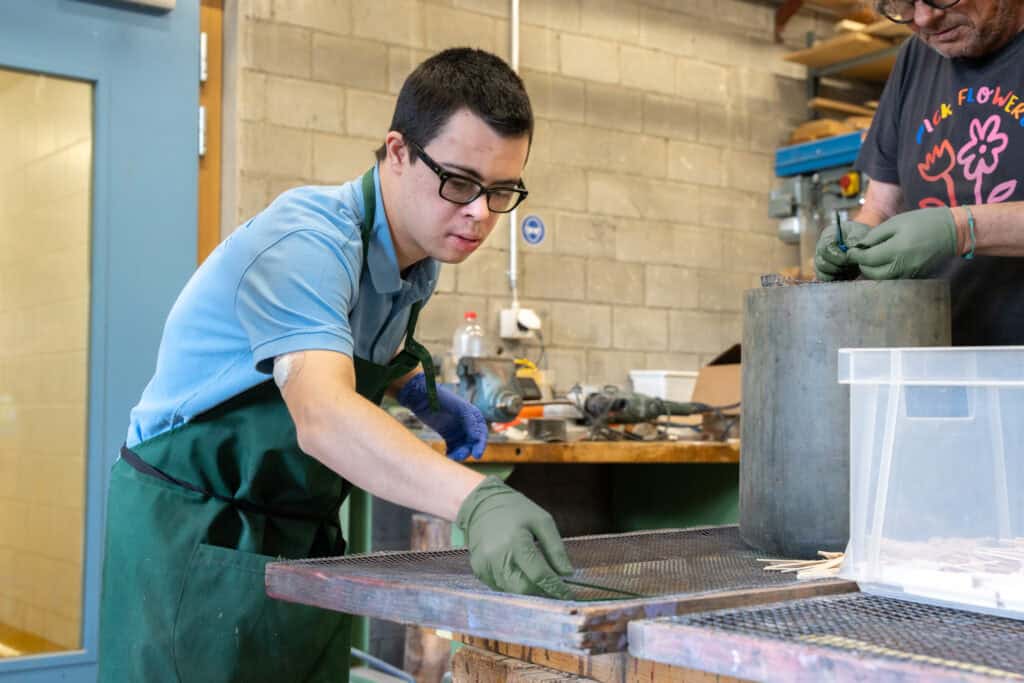 Ein Mann mit Brille und grüner Schürze arbeitet in einer Werkstatt und sortiert oder ordnet mit behandschuhten Händen Gegenstände auf einem Tisch. Eine weitere Person, die ebenfalls Handschuhe trägt und in der Nähe steht, ist teilweise zu sehen. Die Umgebung ist industriell, mit verschiedenen Werkzeugen und Materialien.