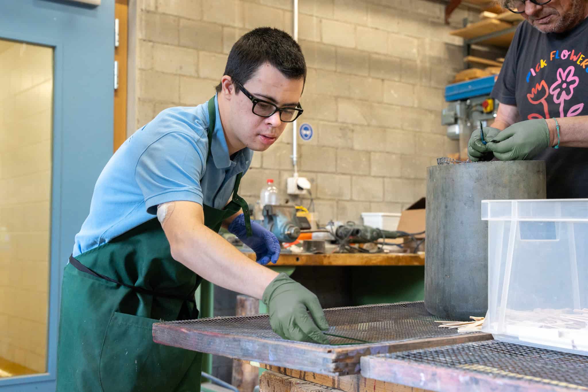Ein Mann mit Brille und grüner Schürze arbeitet in einer Werkstatt und sortiert oder ordnet mit behandschuhten Händen Gegenstände auf einem Tisch. Eine weitere Person, die ebenfalls Handschuhe trägt und in der Nähe steht, ist teilweise zu sehen. Die Umgebung ist industriell, mit verschiedenen Werkzeugen und Materialien.