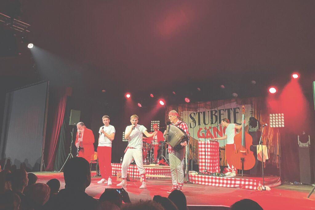 Eine Band tritt unter rotem Licht auf der Bühne auf. Die Gruppe besteht aus einem Sänger, einem Akkordeonspieler, einem Bassisten und einem Bläser. Im Hintergrund steht „Stubete Gäng“. Das Publikum ist im Vordergrund teilweise sichtbar.