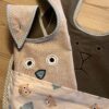 Zwei Esslätzchen-Stofflätzchen mit Tiergesichtern: eines braun mit Ohren und ein beige mit Eulenaugen und -nase, auf einer Holzoberfläche platziert.