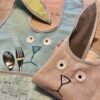Esslätzchen mit Tiermotiven, Lätzchen und Besteck auf einem Holztisch ausgestellt.