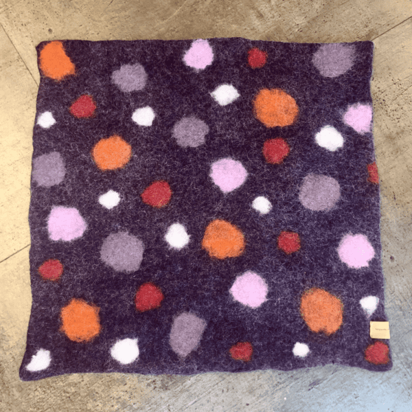 Ein Sitzkissen mit einem lila Stoffquadrat, verziert mit lebendigen kreisförmigen Mustern in Orange, Rosa, Weiß und Rot, auf einer Holzoberfläche platziert.