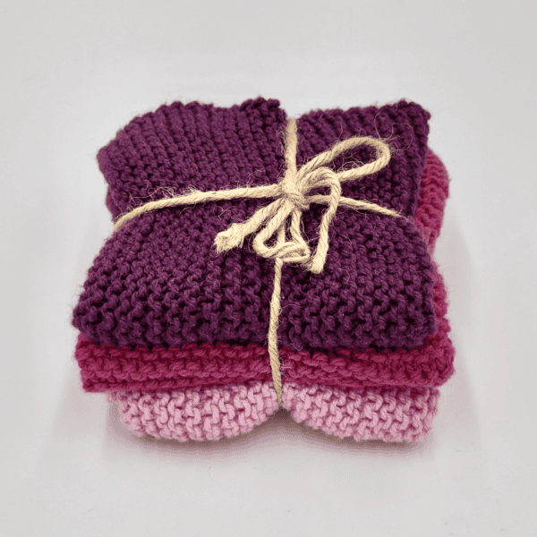 Auf weißem Untergrund liegt ein Stapel gestrickter Spüllappen in Lila und Rosa, zusammengebunden mit einer beigen Kordel.