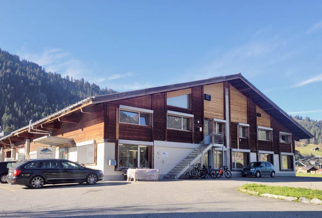 Ein Holzgebäude im Chalet-Stil mit großen Fenstern steht vor einer Bergkulisse. Zwei Autos und einige Fahrräder sind davor geparkt. Der Himmel ist klar und blau und unterstreicht die ruhige Bergkulisse.