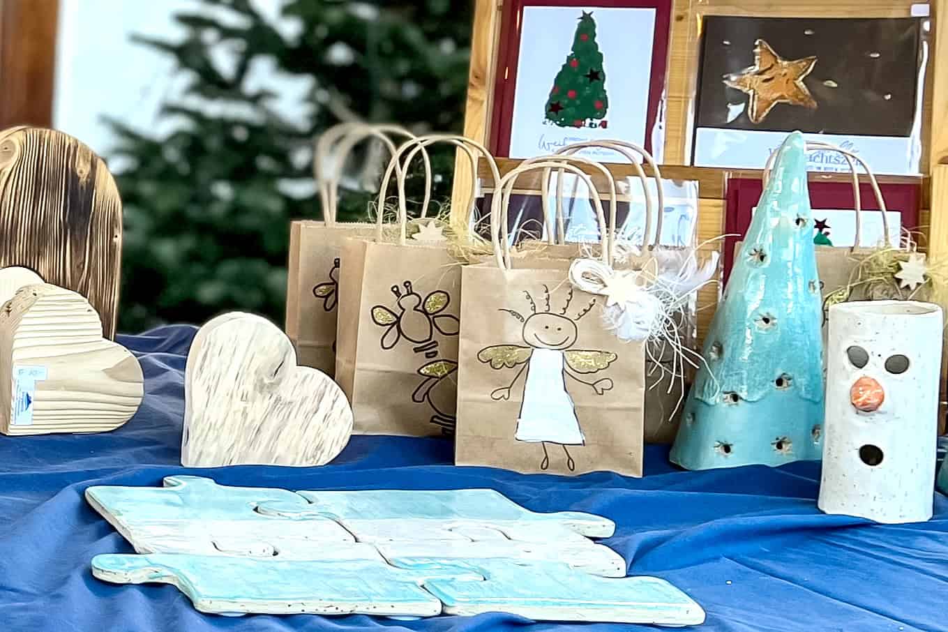 Auf einem Tisch sind handgefertigte Kunsthandwerksgegenstände ausgestellt, darunter Holzherzen, Christbaumschmuck aus Keramik, Geschenktüten aus Papier mit Engel- und Bienenmotiven sowie Grußkarten mit Weihnachtsbaum- und Sternmotiven. Im Hintergrund ist ein verschwommener Weihnachtsbaum zu sehen.