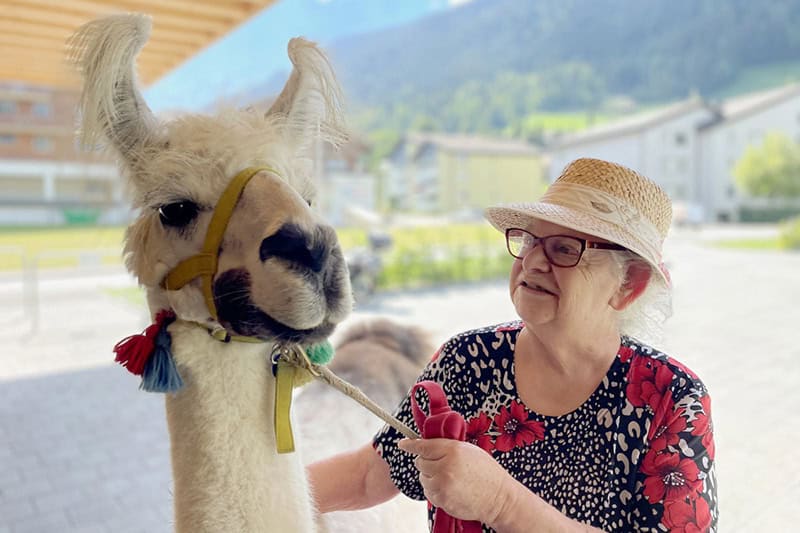 Ältere Frau in Blumenhemd und Strohhut streichelt im Freien ein Lama.