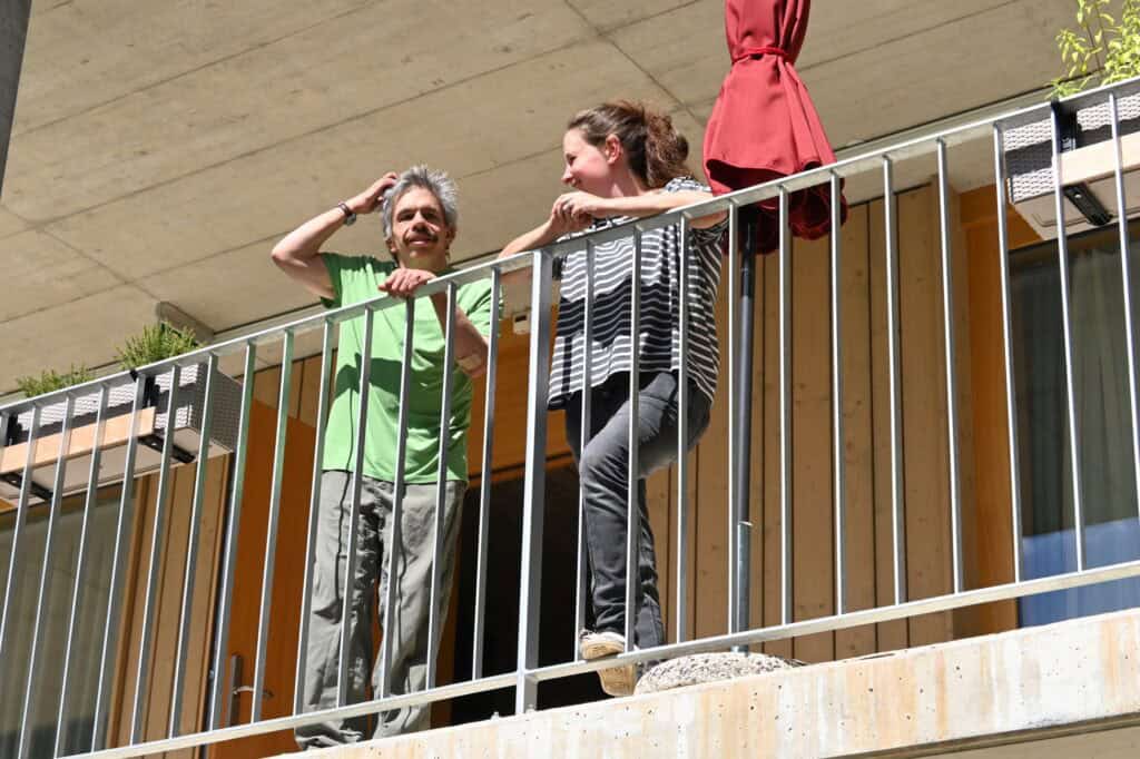 Zwei Menschen unterhalten sich lächelnd auf einem Balkon mit Pflanzen und einem geschlossenen roten Regenschirm.